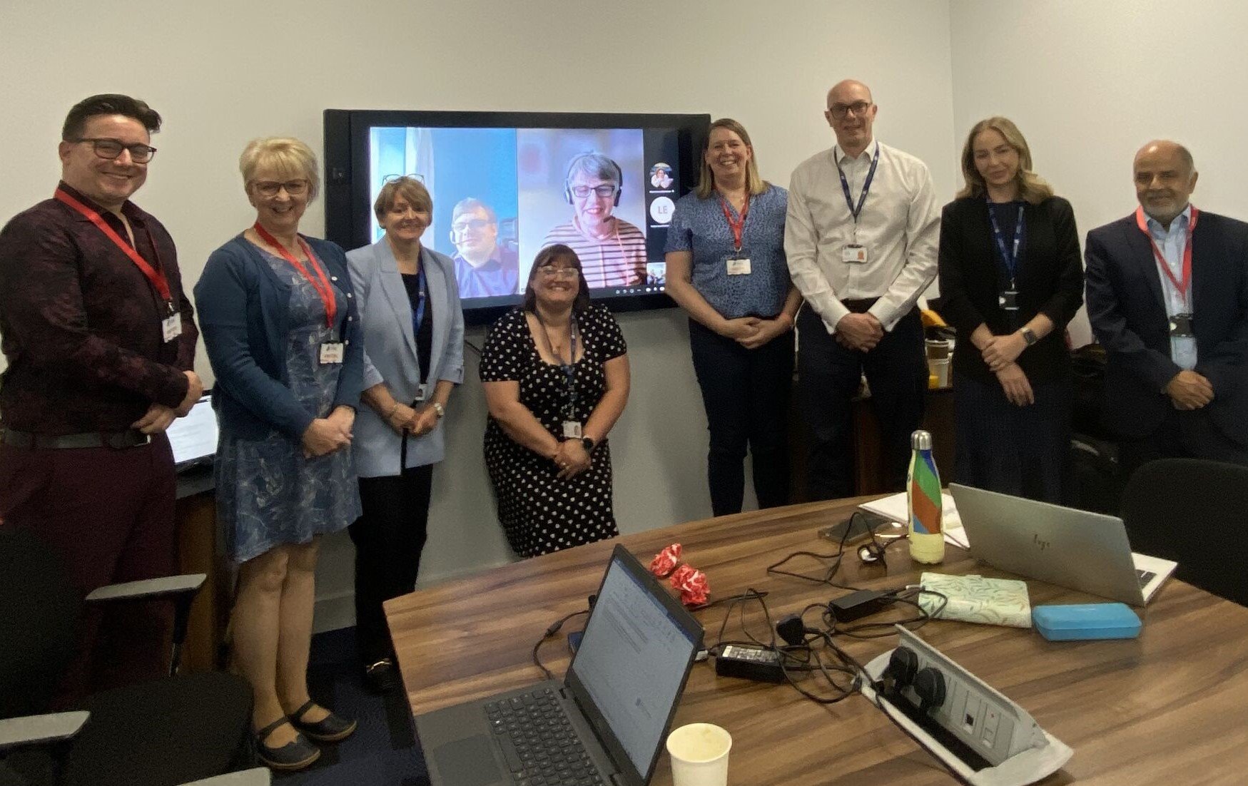 Group of men and women in a meeting room with colleagues on screen in background