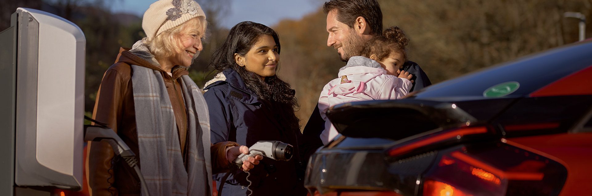 From left to right: Elderly mother charging her electric vehicle, wife, and husband holding child. 